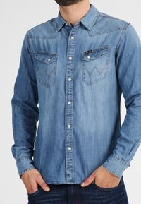 Chemise en denim bleu clair avec manches longues, col pointu, deux poches poitrine, et couture contrastante sur les poches. Fermeture à boutons à l'avant.