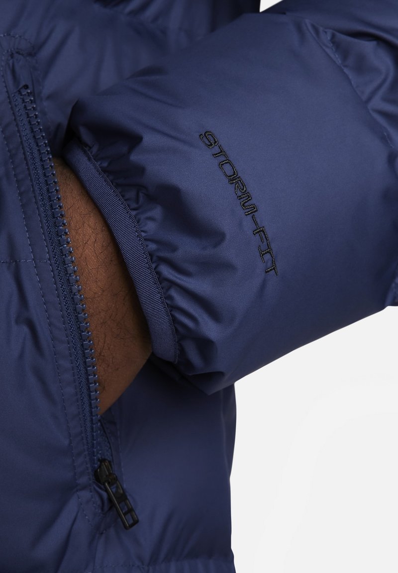 Marineblaue Puffjacke mit glatter Textur, Reißverschluss-Taschen, elastischen Bündchen und gesticktem "STORM-FIT"-Detail am Ärmel.