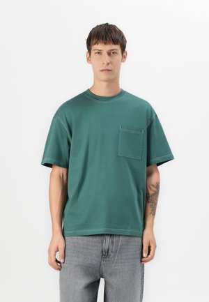 Young man with short brown hair wearing a green pocketed t-shirt and gray jeans, standing against a white background.