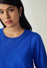 Pull en tricot bleu avec un col rond et de subtiles détails de manches bouffantes. Texture douce, tissu léger.
