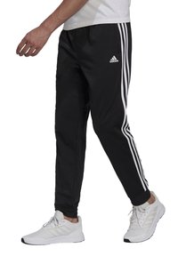 Schwarze Sporthose mit einem weißen Streifendesign in drei Reihen, elastischem Bund und schmalem Beinprofil. Kombiniert mit weißen Sportschuhen.