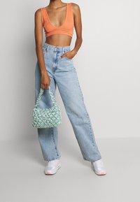 Oranžový crop top, světle modré džíny s vysokým pasem a bílé tenisky. Drží mátově zelenou texturovanou kabelku s řasením.