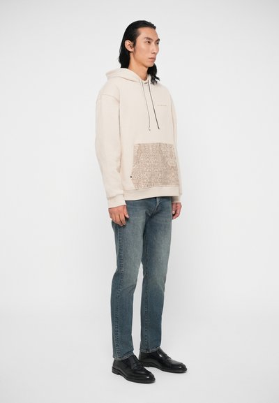 Bežs kapuce ar strukturētu rakstu uz priekšējās kabatas, apvienota ar zilām džinsām un melnām kedām. Gluda auduma un brīva piegriezuma.