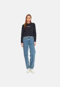 Marineblauwe sweatshirt met het "GUESS" logo, gecombineerd met lichtblauwe ontspannen pasvorm jeans en witte sneakers, staand tegen een effen achtergrond.