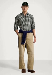 Mann iført svart caps, grønn og hvit rutete skjorte, beige bukser, marineblå genser knyttet rundt livet, og brune sko, står med hendene i lommene.