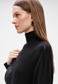 Μαύρο πουλόβερ τουρμπάνι με ριγωτή υφή, που διαθέτει ανοιχτό πλέκτο στα μανίκια, φορεμένο με το χέρι να στηρίζεται στο ύφασμα.