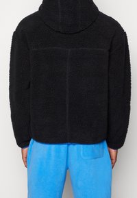 Czarna polarowa kurtka o teksturowanej powierzchni, z kapturem i luźnym krojem. W połączeniu z jasnoniebieskimi spodniami.