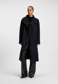 Donna in piedi che indossa un lungo cappotto nero doppiopetto, un top nero a coste e pantaloni neri lucidi a gamba larga su uno sfondo neutro.