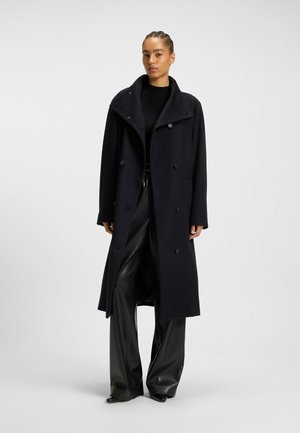 Femme se tenant debout vêtue d'un long manteau noir croisé, d'un top noir à côtes et d'un pantalon noir large et brillant, sur fond uni.