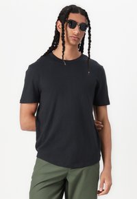 Schwarzes kurzärmliges T-Shirt aus leichtem Stoff, runder Ausschnitt, lockere Passform, kombiniert mit olivgrünen Hosen, Modell trägt schwarze Sonnenbrille.