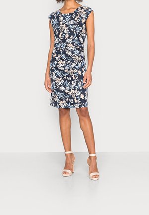 Kvinde iført marineblå blomsterkjole til knæet med kapærmer, kombineret med beige ankelremssandaler med høje hæle, stående på hvidt gulv.