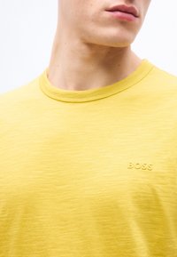 Κοντινό πλάνο ενός ατόμου που φοράει κίτρινη μπλούζα με στρογγυλή λαιμόκοψη και τη λέξη "BOSS" ανάγλυφη στο στήθος.