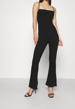 Combinaison noire avec corsage ajusté, jambes évasées et fines bretelles spaghetti. Le matériau semble lisse avec des coutures minimales. Paire de sandales noires à talons.