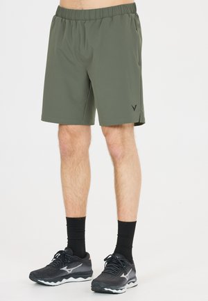 Male wearing olive green athletic shorts, black crew socks, and black running shoes standing on a plain white background.