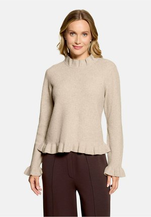 Woman with light brown hair wears beige ribbed sweater with ruffled neck, cuffs, and hem paired with dark brown trousers, standing upright.