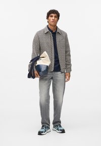 Herre vævet jakke i beige og marineblå, kombineret med en mørkeblå polo, grå jeans og multikolorerede sneakers. Holder en tofarvet taske.