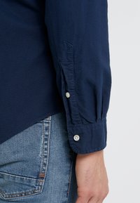 Námořnická modrá košile s knoflíky a texturovanou tkaninou, se zaoblenými manžetami a stříbrnými knoflíky, doplněná o světlé modré džíny.