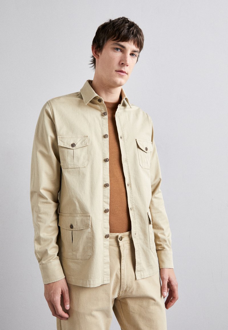 Beige cotton shirt jacket with button front, two chest pockets, and side pockets. Worn over a brown sweater, textured fabric.