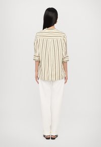 Striped button-up shirt in cream and brown with rolled sleeves, paired with loose white trousers and flat brown shoes. Back view.