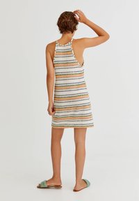 Gestreiftes Halterneck-Kleid mit bunten horizontalen Linien. Weicher, strukturierter Stoff. Offenes Rückendesign. Getragen mit grünen Rutschsandalen.