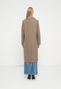 Manteau long en laine marron avec une silhouette droite, col cranté et poches latérales, associé à un pantalon en jean bleu à jambes larges et des talons noirs.