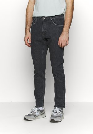 Mies, jolla on mustat kapeat farkut, valkoiset sukat, harmaat ja valkoiset lenkkarit sekä valkoinen t-paita, joka on työnnetty housuihin, seisomassa valkoista taustaa vasten.