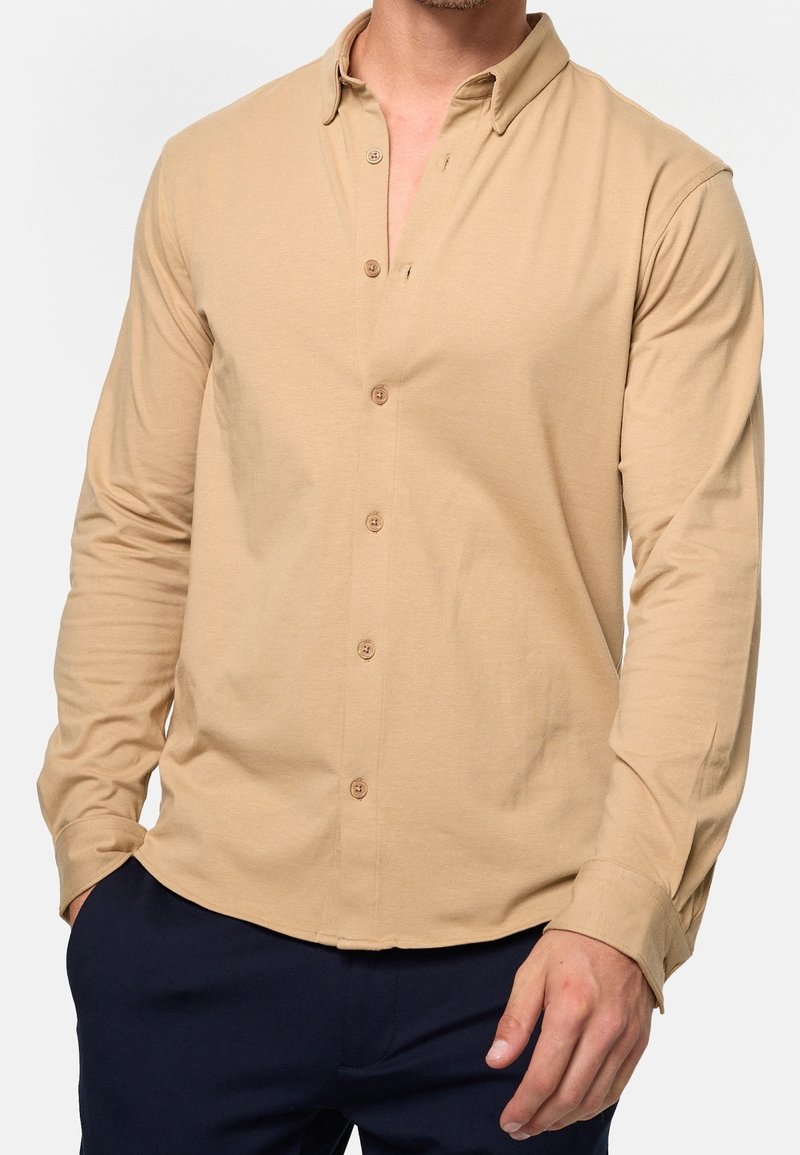 Homme portant une chemise beige à manches longues avec col et boutons, associée à un pantalon bleu marine foncé, debout avec une main dans la poche.