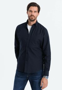 Camisa de botones azul marino de mezcla de algodón con mangas largas y un bolsillo en el pecho, combinada con jeans oscuros y un reloj plateado.