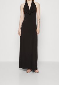 Halter neck black maxi dress made of smooth fabric, featuring a draped front, and a fitted silhouette, paired with clear heeled sandals.