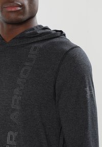 Ciemnoszara bluza z kapturem o teksturowanym wykończeniu, z wyraźnym logo z materiału odblaskowego z lewej strony i na prawym rękawie.