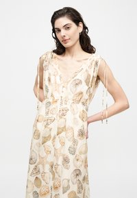 Γυναίκα με μπεζ φόρεμα χωρίς μανίκια, με εκτύπωση κοχυλιών και αστερία, V-λαιμόκοψη και λεπτομέρειες δέσιμου στους ώμους, ποζάρει με το ένα χέρι στη μέση.