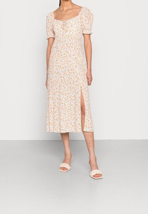 Kvinde iført en hvid midi-kjole med orange blomsterprint, pufærmer, firkantet udskæring, frontslids og flettede hvide sandaletter med åbne tæer.