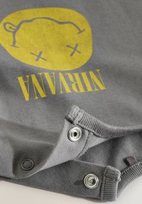 Chemise en coton grise avec un graphique circulaire jaune et du texte à l'envers disant "NIRVANA". Comprend des fermetures à pression métalliques au niveau de l'ourlet.