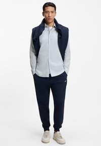 Αθλητικό σύνολο σε ναυτικό μπλε χρώμα που περιλαμβάνει ένα πουλόβερ περασμένο πάνω από ένα ανοιχτό μπλε πουκάμισο με κουμπιά, συνδυασμένο με τζόγκερ ίδιου χρώματος και μπεζ αθλητικά παπούτσια.