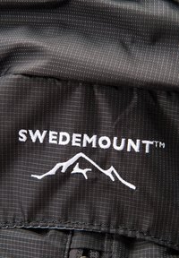 Tissu noir texturé avec le logo "SWEDEMOUNT™" en broderie blanche et un graphisme de montagne. Présente une fermeture éclair en bas.