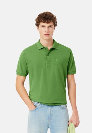 Jeune homme aux cheveux bouclés portant un polo vert et un jean bleu clair, tenant un vêtement vert plié dans sa main droite.