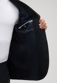 Zwarte blazer met een gladde buitenkant en ingewikkelde voering. Voorzien van een zak met een driehoekige klep, contrasterende stiksels en minimale merkvermelding.