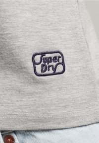 Světle šedé tričko s modrým vyšívaným logem "Super Dry". Látka je měkká s hladkým povrchem a design je jednoduchý a ležérní.
