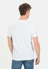 Hvid bomulds t-shirt med kortærmer, rund hals og en ensfarvet ryg; bæres med blå denim jeans og synlig tatovering på venstre arm.