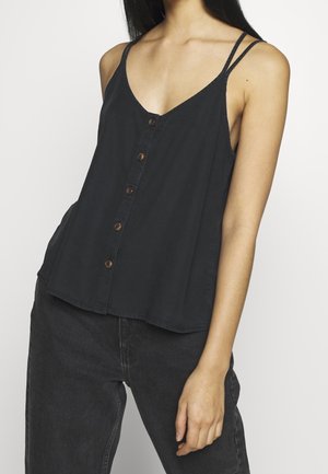 Kobieta ubrana w czarny bezrękawnik na guziki z podwójnymi cienkimi ramiączkami i wysokim stanem czarne spodnie, stojąca na jednolitym tle.