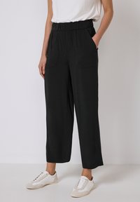Pantalon large noir avec une taille élastique et deux poches avant, fabriqué en tissu doux, associé à des chaussures de sport blanches.