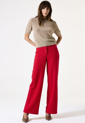 Un pull court à manches courtes en beige et duveteux associé à un pantalon large rouge texturé taille haute et des escarpins métalliques à bout pointu.