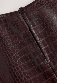 Burgundy faux leather skirt with a crocodile texture, featuring a central zipper closure and a round button accent at the top.