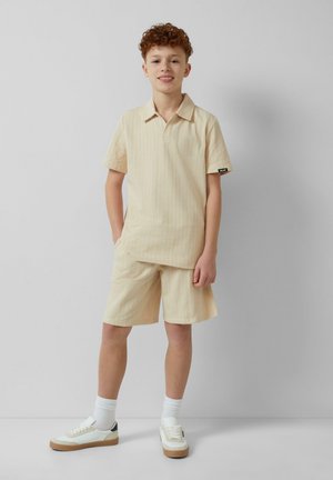 Lichtbeige gestreepte polo met korte mouwen en bijpassende shorts, vervaardigd uit een gestructureerd materiaal, gecombineerd met witte sneakers en witte gestreepte sokken.