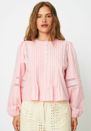 Nő világos rózsaszín hosszú ujjú blúzban, függőleges csíkokkal és csipkebetétekkel, krémszínű, szemmintás szoknyával párosítva.