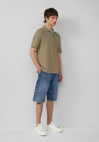 Polo de color verde oliva con mangas cortas y tejido texturizado, combinado con unos pantalones cortos de mezclilla hasta la rodilla con diseño de patchwork y zapatillas blancas.