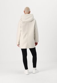 Lys grå ullblandingsfrakk med stor hette, tettsittende ermer og rett silhuett, kombinert med svarte bukser og hvite joggesko.