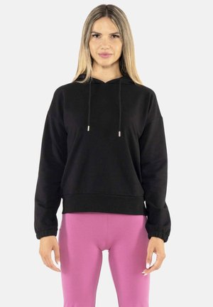 Femme aux cheveux blonds et raides portant un sweat à capuche noir et des leggings roses, debout devant un fond blanc uni.