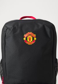 Svart ryggsekk med rød glidelås og håndtak, med en fremtredende sirkulær Manchester United-logo i gult og rødt på forsiden.