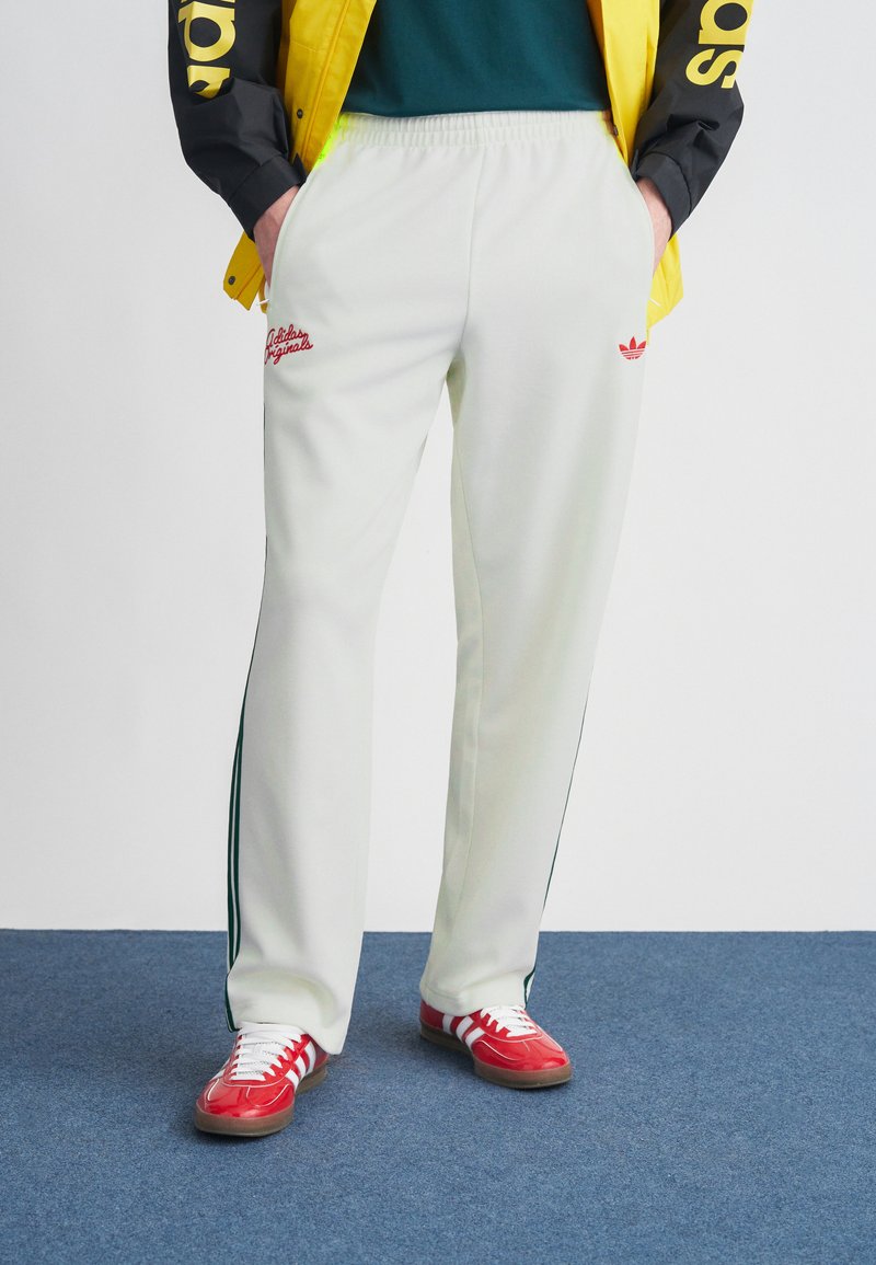 Λευκό αθλητικό παντελόνι με πράσινες ρίγες στο πλάι, με κόκκινο κεντημένο λογότυπο. Συνδυάζεται με κόκκινα αθλητικά παπούτσια με λευκές ρίγες.
